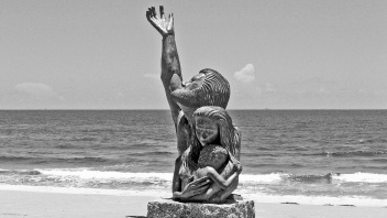 Steinfigur am Meer: Ein Mann hält schützend eine Frau und ein Kind und streckt die andere Hand zum Himmel - Das Wort zum Sonntag