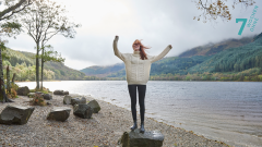 Frau steht mit erhobenen Händen auf einem Stein vor einem See.