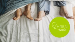 Zwei Paar Füße von Senioren schauen am Fußende eines Bettes unter der Bettdecke hervor