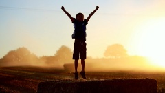 Ein Junge springt in der Abendsonne mit hochgestreckten Armen auf einem Strohballen.