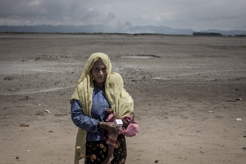Eine Rohingya-Frau mit Kopftuch und Kind auf der Schulter steht in einer öden Ebene und blickt in die Kamera.