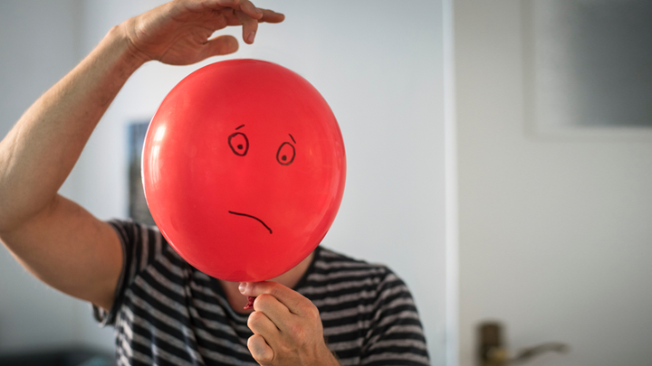 Ein Mann hält einen roten Luftballon, auf den trauriges Gesicht gemalt ist, vor seinen Kopf.