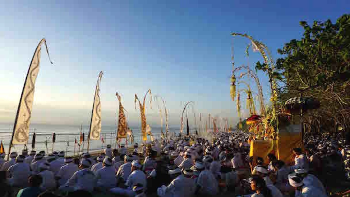 Menschen in festlichen Gewändern vor goldenen Altären und Tempelfahnen am Strand von Bali