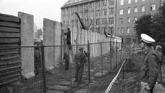 Ein Bautrupp verspachtelt die neu errichteten Betonsegmente, während ein Kran die Rohrauflage auf die neue „Grenzmauer 75“ setzt.